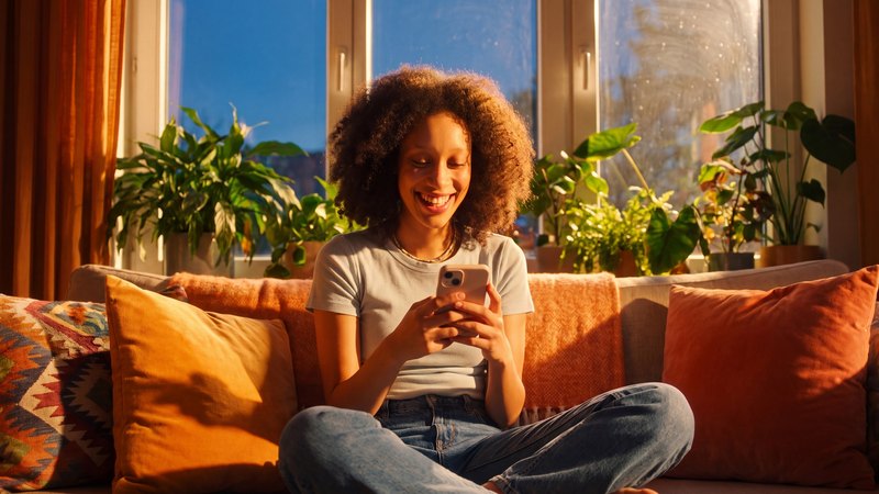 Een creator zit met haar telefoon op de bank bij een zonnig raam met planten