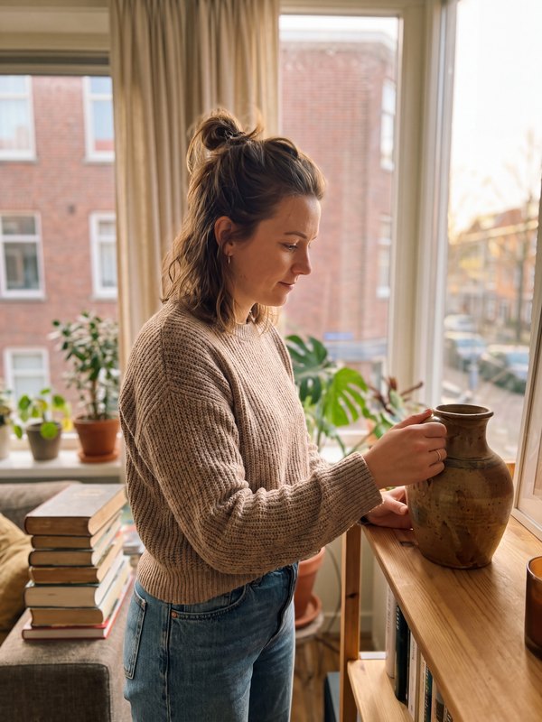 Iemand zet een keramieken vaas op een plank bij het raam, planten en boeken ernaast